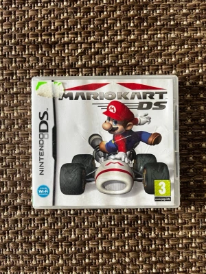 Mario Kart DS till Nintendo DS - Mario Kart DS är ett racingspel till Nintendo DS där du tävlar som Mario och andra karaktärer i färgglada gokarts. Omslaget är vitt med Mario i röd keps och blå overall. Spelet har stöd för Wi-Fi och är PEGI 3-märkt.