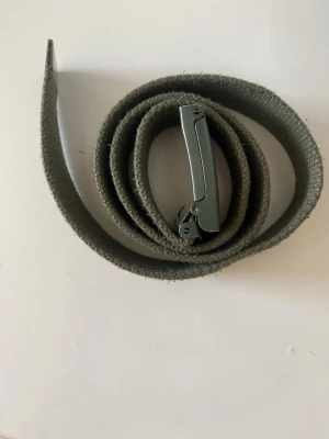 Grönt tygbälte med metallspänne - Snyggt militärgrönt bälte i tyg med ett stilrent metallspänne. Perfekt för att ge en casual touch till din outfit. Bältet är enkelt att justera och passar de flesta stilar.