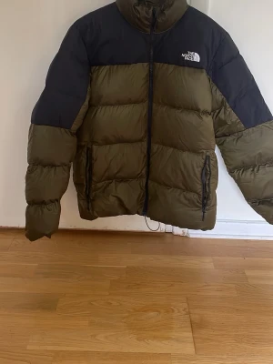 The North Face grön pufferjacka - Säljer en fet pufferjacka från The North Face i olivgrönt med svarta axelpartier och hög krage. Jackan har dragkedja framtill, två sidofickor med dragkedja och klassisk logga på bröstet. Perfekt för kalla dagar och riktigt snygg streetstil.