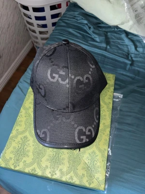 Svart keps med GG-mönster från Gucci - Snygg svart keps från Gucci med det klassiska GG-mönstret över hela kepsen. Kepsen har böjd skärm och är tillverkad i ett slitstarkt tyg med detaljer i svart. Perfekt för dig som vill ha en stilren accessoar med lyxig känsla. Pris går att diskuteras.