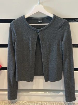 Mörkgrå kofta från Gina Tricot - Mörkgrå, tunn och mjuk kofta från Gina Tricot med rund halsringning och en knapp framtill. Kortare modell med långa ärmar, perfekt att slänga över en topp. Enkel och stilren design som passar till det mesta.Lite noprig men inget som märks jätte mycket.