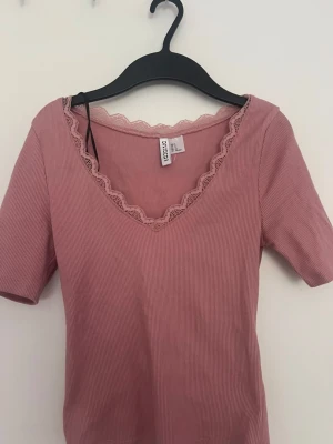 Rosa ribbad topp med spets från COSMO - Supersöt ljusrosa ribbad topp från COSMO med v-ringning och spetsdetaljer längs halsen. Kortärmad och figurnära modell som är perfekt till jeans eller kjol. Materialet känns mjukt och stretchigt.