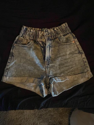 Högmidjade denimshorts med resår - Säljer ett par ljusblå högmidjade jeansshorts med resår i midjan och uppvikta benslut. Shortsen har klassiska fem fickor och stängs med dragkedja och knapp. Perfekta för sommaren och ger en avslappnad vibe.