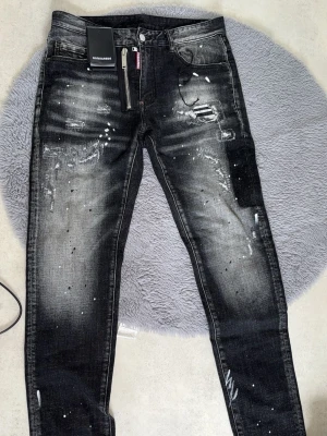 Svarta Dsquared2 jeans med slitningar - Coola svarta jeans från Dsquared2 med slitningar, färgstänk och detaljerad dragkedja framtill. Jeansen har en slim passform och är tillverkade i jeansmaterial med snyggt tvättad look och unika detaljer. Perfekta för dig som vill sticka ut med din stil.