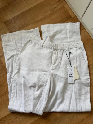 Vita linnebyxor från Gina Tricot  - Snygga vita linnebyxor från Gina Tricot med raka ben och klassisk passform. Byxorna har fickor på sidorna, hällor för bälte och är tillverkade i 100% linne för en luftig och skön känsla. Perfekta för sommarens alla dagar.