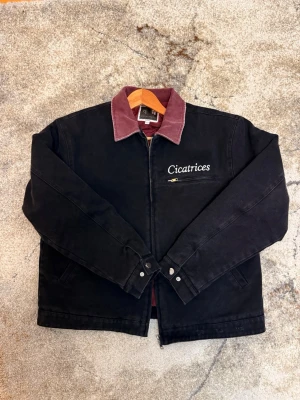 Workwear Cicatrices - Stilren svart jacka med dragkedja från lowfrms. Jackan har en kontrasterande vinröd krage i manchestertyg och broderad logga på bröstet. Två sidfickor och en bröstficka med dragkedja. Perfekt för lager-på-lager och streetwear.