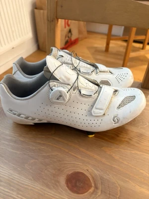 Vita cykelskor från Scott - Säljer ett par vita cykelskor från Scott med BOA-snörning och kardborreband för perfekt passform. Skorna har ventilerande meshpaneler och perforerat material för bra andningsförmåga. Snygg och sportig design, perfekta för landsvägscykling.