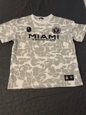 BAPE x Inter Miami camo t-shirt - Snygg kortärmad t-shirt från BAPE i samarbete med Inter Miami. T-shirten har grått och vitt camouflagemönster, tryck med MIAMI och A BATHING APE på bröstet samt klubbmärke och BAPE-logga. Tillverkad i mjuk bomull och har normal passform.