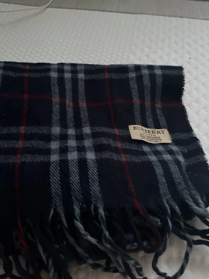Mörkblå rutig halsduk från Burberry - Snygg mörkblå halsduk från Burberry med klassiskt rutmönster i grått, vitt och rött. Halsduken har tvinnade fransar i kanterna och en tygpatch med Burberry-loggan. Perfekt accessoar för att lyfta din outfit.