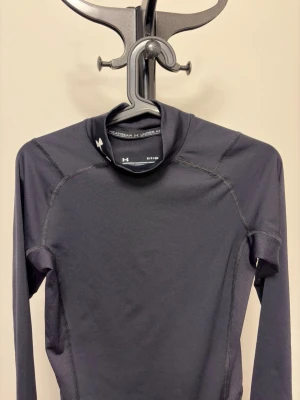 Under Armour Turtle Neck Compression shirt - Snygg svart långärmad träningströja från Under Armour med tight passform och diskret logga vid halsen. Tillverkad i ett stretchigt funktionsmaterial som andas, perfekt för intensiva träningspass eller löpning. Sömmen är markerad för extra stil och komfort.