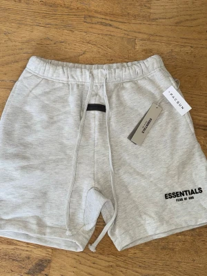 Grå Essentials Fear of God shorts - Snygga ljusgrå shorts från Fear of God Essentials med svart logga på benet. De har elastisk midja med snörning och är gjorda i mjuk bomull, perfekta för en chill och avslappnad stil. Kommer med originaltagg från Pacsun.
