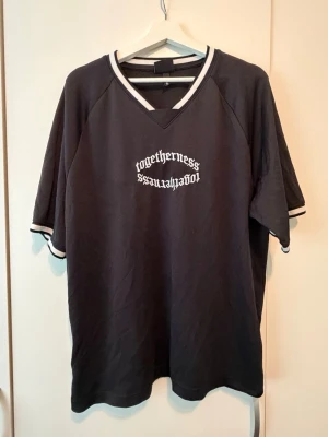 Svart v-ringad t-shirt med tryck - Svart t-shirt i bomull med vit text i gotisk stil på bröstet. V-ringad hals med vit kant och matchande vita detaljer på ärmsluten. T-shirten har loose passform och korta ärmar, perfekt för en avslappnad streetlook.