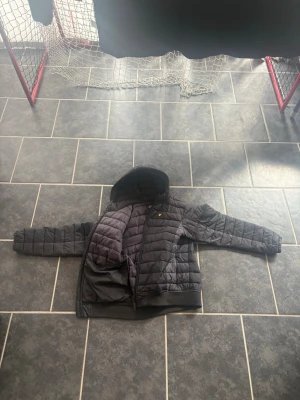 Svart pufferjacka med huva Lyle & Scott - Säljer en svart pufferjacka från Lyle & Scott med huva och dragkedja. Jackan har quiltad design, ribbade muddar och den klassiska loggan på bröstet. Perfekt för kalla vinterdagar och riktigt skön att bära. Materialet är syntet och håller värmen bra.