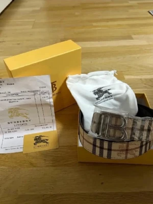 Burberry rutigt bälte med stor B-spänne - Säljer ett klassiskt Burberry bälte med det ikoniska rutiga mönstret i beige, svart och rött. Bältet har ett stort silvrigt B-spänne framtill och kommer med originalpåse och box. Perfekt accessoar för att lyfta din outfit.