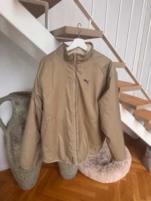 Beige vindjacka från Puma - Stilren beige vindjacka från Puma med dragkedja framtill och hög krage. Jackan har en minimalistisk look med Puma-logga på bröstet och är tillverkad i ett lätt, vindtätt material. Perfekt för blåsiga dagar och enkel att matcha med olika outfits.