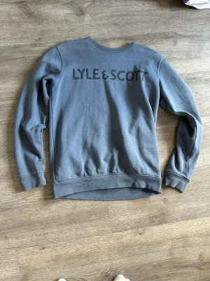 Blå sweatshirt från Lyle & Scott - Snygg blå sweatshirt från Lyle & Scott Junior med stor logga på ryggen. Tröjan har rund halsringning, långa ärmar och ribbade muddar vid ärmslut och nederkant. Perfekt för en avslappnad och trendig look.