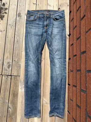 Blå slim fit jeans från Nudie Jeans - Säljer ett par klassiska blå slim fit jeans från Nudie Jeans med snygg slitning och kontrastsömmar. Jeansen har fem fickor, knappgylf och är tillverkade i denim med en skön stretch. Perfekta till en avslappnad stil.