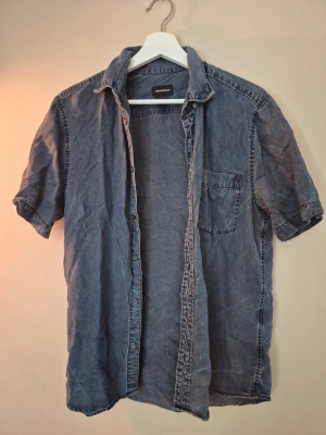 Blå kortärmad skjorta H&M Conscious - Säljer en blå kortärmad skjorta från H&M Conscious. Skjortan har klassisk krage, bröstficka och knappar framtill. Tillverkad i mjuk bomull och har en avslappnad passform som passar perfekt till linnébyxor eller shorts.