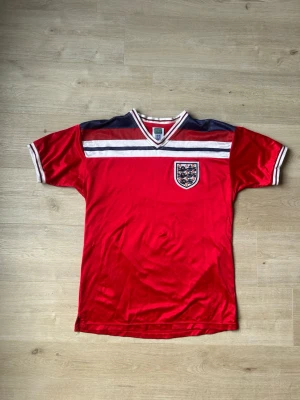 Röd England Retro Fotbollströja Storlek L - Mycket fin fotbollströja i röd färg. Tröjan är i mycket bra skick och sparsamt använda. |  Äkta som allt annat jag säljer! ✅ |  Storlek: L | Skick: Mycket Bra Skick -  (SE SISTA BILD)| Färg: Röd | Märke: England | Modell: Fotbollströja |   Hör gärna av dig vid frågor så svarar jag så fort jag kan! 😁