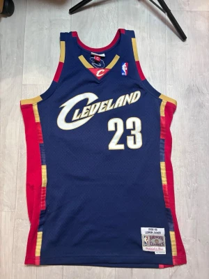 Cleveland Cavaliers basketlinne #23 - Snyggt basketlinne från Mitchell & Ness med Cleveland Cavaliers tryck och nummer 23. Marinblå bas med röda och gula detaljer, samt 'James' och '23' på ryggen. Tillverkat i ventilerande meshmaterial, perfekt för basket eller streetwear.