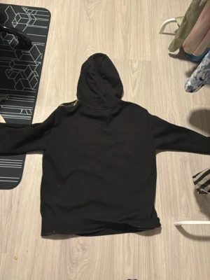 Svart EA7 Emporio Armani hoodie - Snygg svart hoodie från EA7 Emporio Armani med guldfärgad logga på bröstet och detaljer på ärmen. Klassisk huva med dragsko och stor magficka framtill. Tillverkad i mjukt material som känns skönt mot huden. Perfekt för en avslappnad och trendig look.