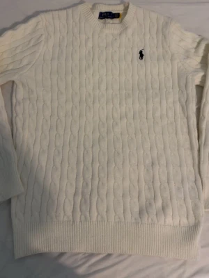 Vit kabelstickad tröja Polo Ralph Lauren - Kabelstickad vit tröja från Polo Ralph Lauren med rund halsringning och broderad logga på bröstet. Tröjan har långa ärmar och ribbade muddar vid ärmslut och nederkant. Klassisk och stilren design i mjukt material.