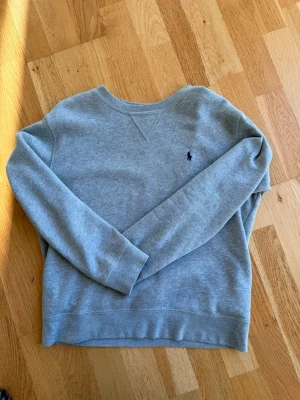 Grå sweatshirt från Polo Ralph Lauren - Klassisk grå sweatshirt från Polo Ralph Lauren med rund halsringning och broderad marinblå logga på bröstet. Tröjan har långa ärmar och är gjord i mjukt bomullsmaterial för en avslappnad och clean look. Tröjan är storlek M för barn ca 10-12 år.