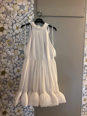 Vit kortklänning från Malina - Liana halterneck mini dress är en söt vit kortklänning från Malina med volangkant nedtill och hög hals. Klänningen har ärmlös design och knytband i nacken för extra detalj. Storlek L - nypris 2999kr