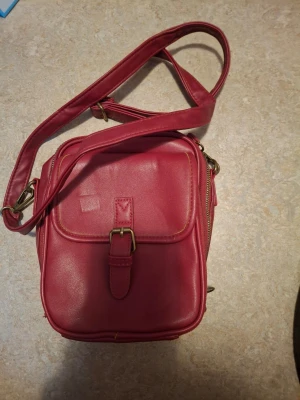 Mörkröd axelväska i skinnimitation - Snygg mörkröd axelväska i skinnimitation med justerbar axelrem och guldfärgade metalldetaljer. Väskan har ett framfack med spänne och dragkedja upptill. Perfekt storlek för det viktigaste och enkel att matcha till olika outfits.