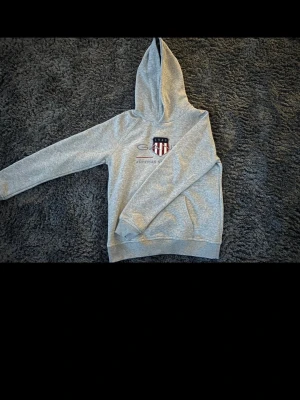 Gant hoodie - Snygg grå hoodie från Gant med huva och ett broderat amerikanskt sköldmärke på bröstet. Klassisk passform med ribbade muddar vid ärmslut och nederkant. Perfekt för en avslappnad och sportig stil. Mjuk bomullsblandning för extra komfort.