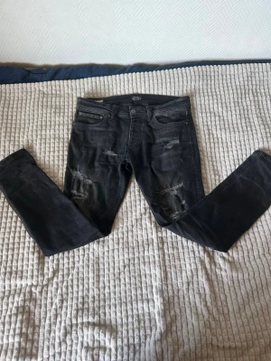 Jack and Jones W32 passar W31  - Säljer ett par svarta jeans från Jack and Jones med slitningar och trasiga detaljer på både fram- och baksida. Jeansen har klassisk femficksmodell och är tillverkade i stretchigt denimtyg för en tajt passform. Perfekta för en edgy och avslappnad stil.
