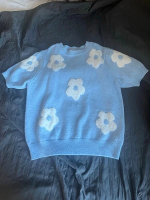 Blå stickad tröja med blommor - Supersöt ljusblå stickad tröja med stora vita blommor. Tröjan har korta ärmar och rund halsringning, perfekt för dig som gillar en lekfull och trendig stil. Passar till både jeans och kjol för en chill look. Vet ej varumärke💓i storlek S, är använd en gång💓💓