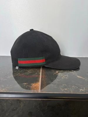 Svart keps från Gucci med GG-mönster - Snygg svart keps från Gucci med diskret GG-monogram över hela tyget. På sidan finns den klassiska gröna och röda randen som ger en exklusiv touch. Kepsen har böjd skärm och är tillverkad i ett slitstarkt tyg, perfekt för dig som gillar streetwear och designeraccessoarer.