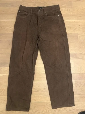 Bruna manchesterbyxor från H&M - Säljer ett par bruna manchesterbyxor från H&M med klassisk femficksmodell och raka ben. Byxorna har synliga sömmar och silverfärgade nitar vid fickorna. Perfekta för dig som gillar retrostil och mjukt material.