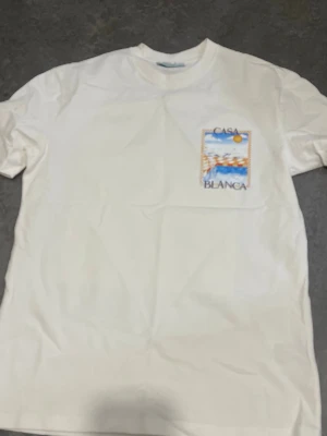 Vit t-shirt från Casablanca - Vit t-shirt från Casablanca med färgglad tryckt patch på bröstet som visar ett soligt landskap och texten 'Casa Blanca'. Klassisk passform och rund hals. Perfekt för dig som gillar stilrena plagg med en twist.