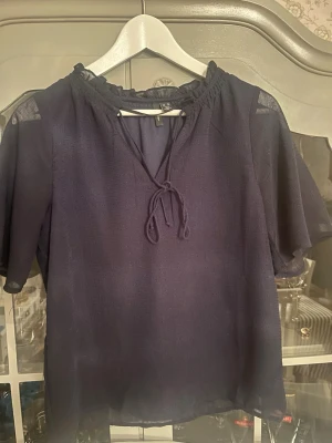 Mörkblå blus med knyt från Vero Moda - Snygg mörkblå blus från Vero Moda med korta, vida ärmar och knytband vid halsen. Blusen har en lätt transparent känsla och volangdetalj vid halsringningen. 