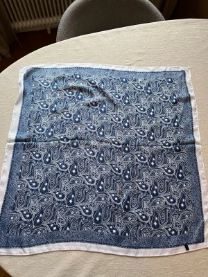Blå paisleymönstrad scarf - Snygg blå scarf med vitt paisleymönster och vit kant. Fyrkantig form som passar perfekt att knyta runt halsen eller i håret. Tillverkad i ett mjukt och lätt material. Märkt 'Fashion Scarf'.