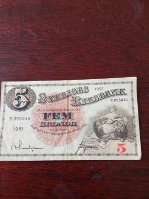 Femkronorssedel Sveriges Riksbank 1951 - Säljer en svensk femkronorssedel från 1951, utgiven av Sveriges Riksbank. Sedeln har dekorativa detaljer, siffran 5 i hörnen och motiv av en kvinna med lejon. Baksidan har porträtt och texten 'Sveriges Riksbank'. Perfekt för samlare av gamla sedlar.