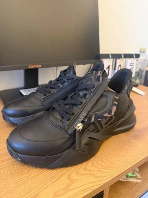 Svarta Fendi sneakers med logga - Snygga svarta sneakers från Fendi med chunky sula och detaljer i svart läder. Skorna har Fendi-logga i camomönster på plösen och hälen samt dragkedjedetalj på sidan. Perfekta för dig som gillar exklusiv streetwear och vill sticka ut.