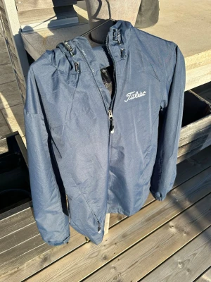 Mörkblå regnjacka från Titleist - Snygg mörkblå regnjacka från Titleist med huva och dragkedja framtill. Jackan har två sidofickor med dragkedja. Perfekt för blöta dagar och enkel att packa ner i väskan. Storlek L men ganska liten i passform