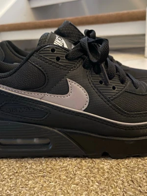 Nike Air Max 90 svart/silver sneakers stl 38,5 - Snygga Nike Air Max 90 sneakers i svart med silverfärgad swoosh. Skorna har en klassisk låg siluett, mesh och syntetmaterial i ovandelen samt den ikoniska Air Max-dämpningen i sulan. Perfekta för dig som gillar streetwear och sportig stil.(Har bara använts en gång, säljs pga felköp).
