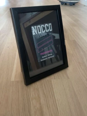 Svart fotoram med NOCCO-bild - Säljer en svart fotoram med en bild av NOCCO Passion, tryckt med vit och rosa text. Ramen är rektangulär och står på ett trägolv. Perfekt som dekoration för dig som gillar energidrycker eller vill ha något unikt på väggen.