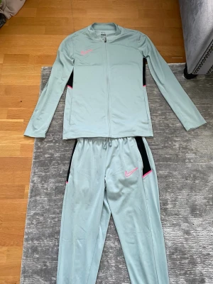 Nike ljusblå träningsset - Nike träningsset i ljusgrått med långärmad zip-jacka och matchande byxor. Setet har svarta och rosa detaljer samt Nike-logga i rosa på både jacka och byxa. Tillverkat i ett stretchigt funktionsmaterial som passar perfekt till träning.