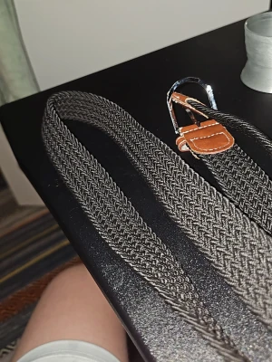 Flätat grått bälte med läderdetaljer - Snyggt flätat bälte i grått med bruna läderdetaljer vid spännet. Bältet har ett silverfärgat metallspänne och är elastiskt för extra komfort. Passar perfekt till jeans eller chinos för en avslappnad men stilren look.