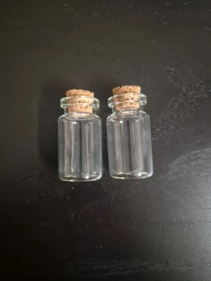 Små glasflaskor med kork - Två genomskinliga små glasflaskor med rund form och korklock. Perfekta för pyssel, dekoration eller förvaring av småsaker. Flaskan är helt transparent och korken är ljusbrun.