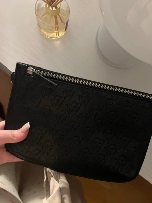 Svart kuvertväska  - Snygg svart kuvertväska från Carin Wester med präglad logotyp över hela väskan. Väskan har silverfärgad dragkedja och är gjord i konstläder. Perfekt att bära i handen för en stilren look.