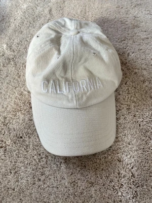 Beige keps med CALIFORNIA-brodyr - Snygg beige keps med böjd skärm och broderad texten 'CALIFORNIA' framtill. Tillverkad i bomullstyg med ventilerande hål upptill för extra komfort. Perfekt accessoar för en avslappnad streetwear-look.