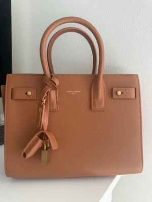 Brun Sac de Jour handväska  - Säljer en brun Sac de Jour handväska  i strukturerat läder. Väskan har två topphandtag, guldfärgade metalldetaljer och ett hänglås med logotyp. Klassisk boxig form och diskret logga framtill.