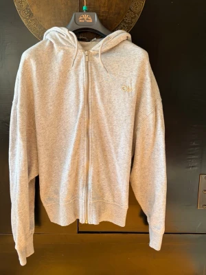 Grå zip hoodie från Carin Wester - Säljer en stilren grå hoodie från Carin Wester med dragkedja framtill och broderad logga på bröstet. Perfekt för en avslappnad och trendig look.