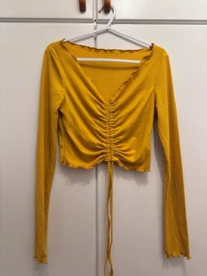 Gul ribbad topp med snörning - Trendig gul långärmad topp med ribbad struktur och djup v-ringning. Toppen har snörning framtill som ger en snygg rynkad effekt och croppad passform. Perfekt för dig som vill sticka ut med färg och coola detaljer.